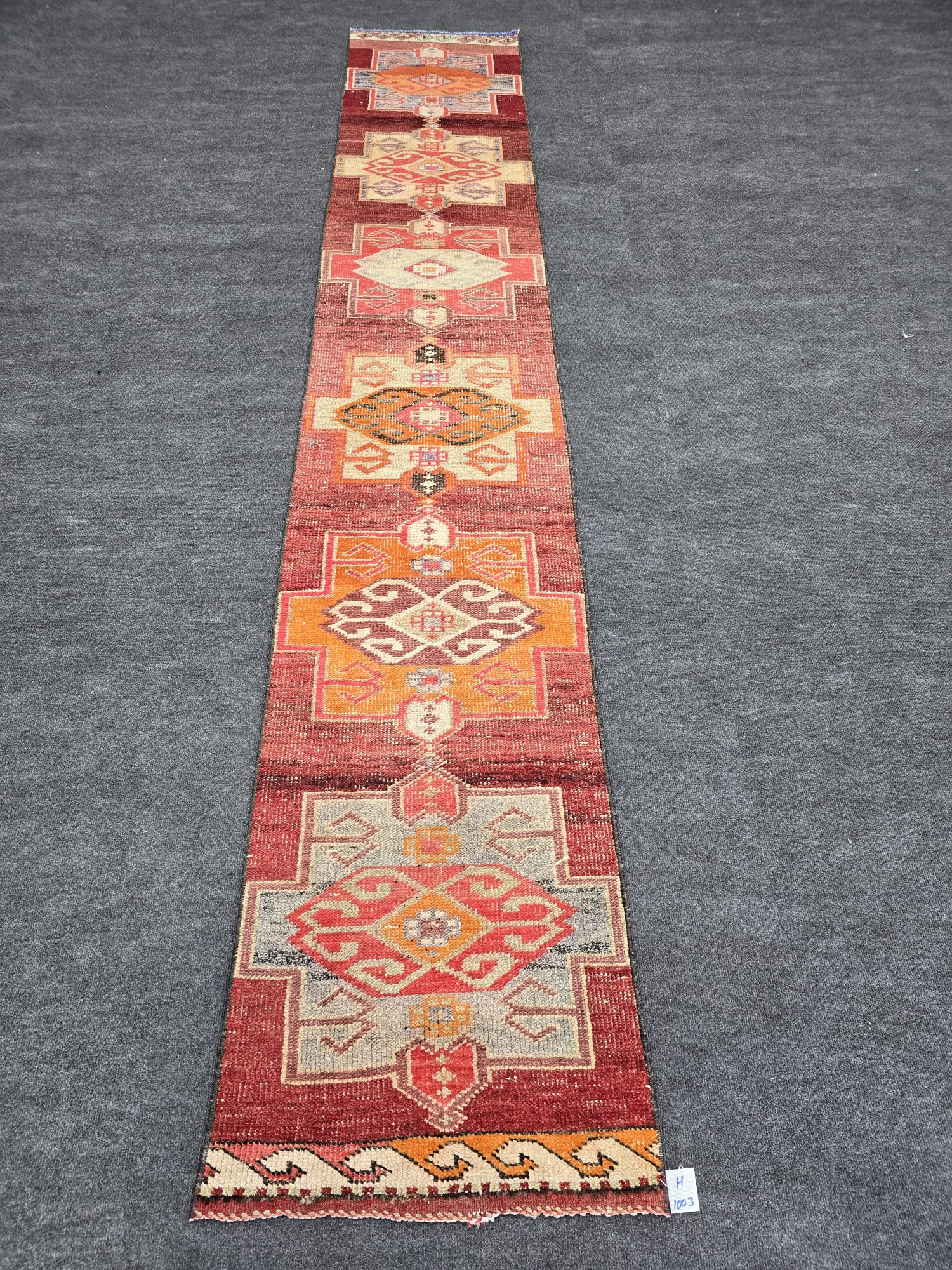 A long, narrow vintage Turkish runner rug featuring a dominant red background with repeating geometric medallions in various shades of orange, beige, and gray. The rug has a distressed appearance and is laid on a dark gray carpet.